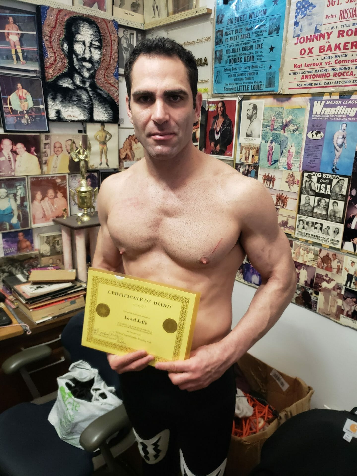 Israel Joffe holding a framed certificate in a gym office covered with wrestling posters